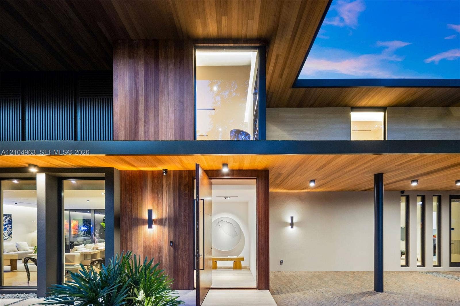 Contemporary facade featuring extensive wood-paneled cladding, floor-to-ceiling glass windows, recessed ceiling lighting, a wide pivot entry door, and covered stone paver driveway