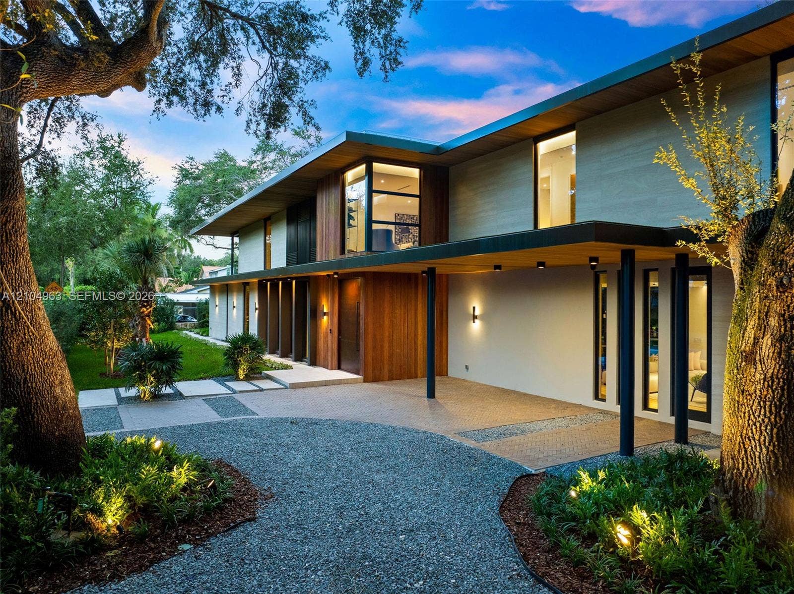 Contemporary architectural exterior featuring a combination of wood-paneled siding and smooth finish surfaces, extensive roof overhangs, custom-designed car shelter, and a multi-textured driveway with integrated paver and gravel pathways