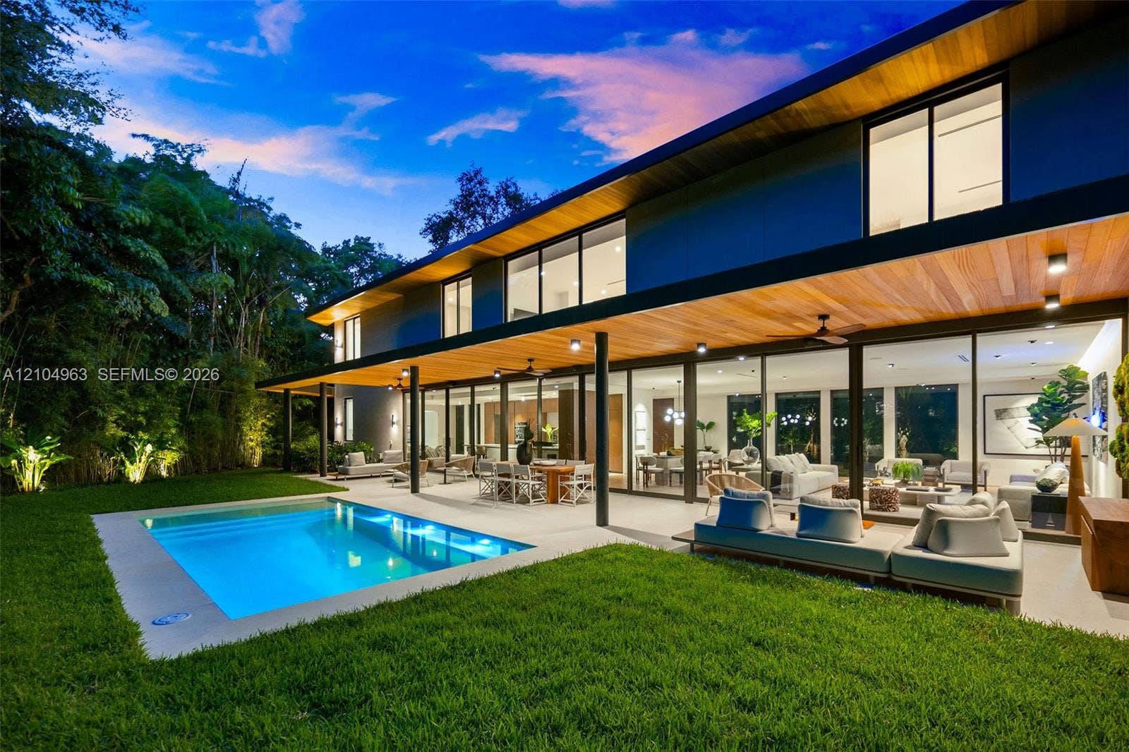 Modern architectural exterior featuring expansive wood-paneled eaves, full-height sliding glass walls, a covered concrete patio, and an in-ground swimming pool set within a manicured lawn