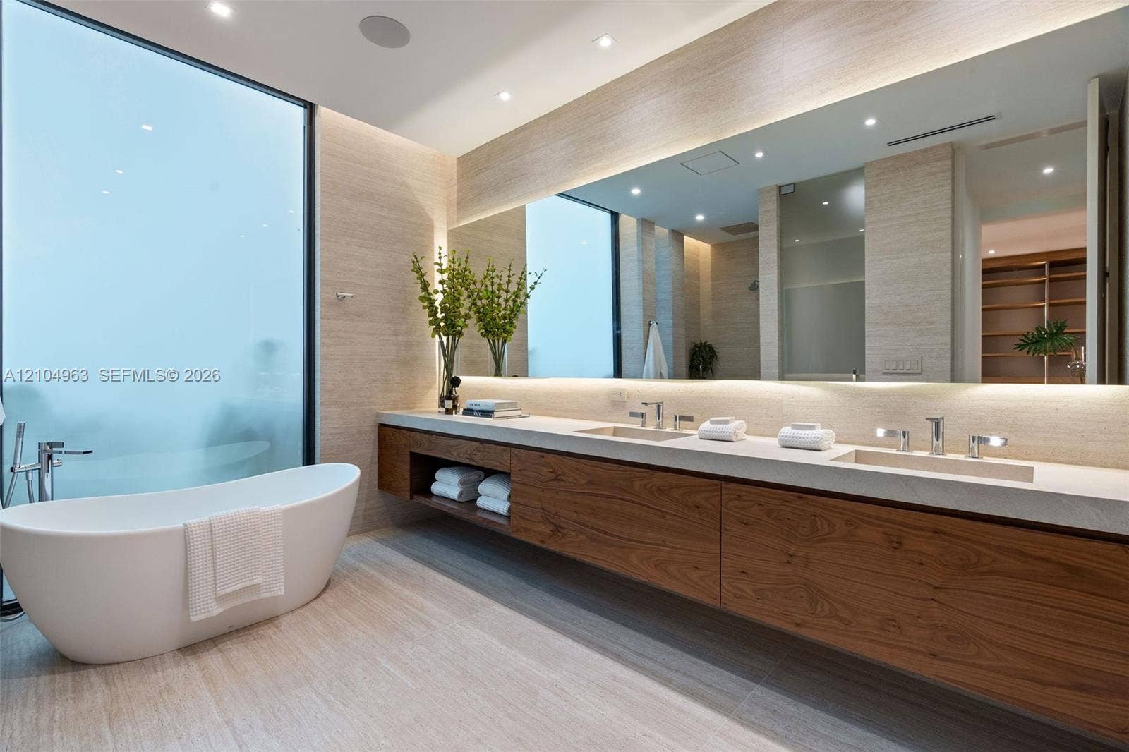 Floating wood-finish vanity with dual integrated sinks, light-toned stone countertop, freestanding soaking tub, expansive wall-to-wall mirror, and recessed ceiling lighting