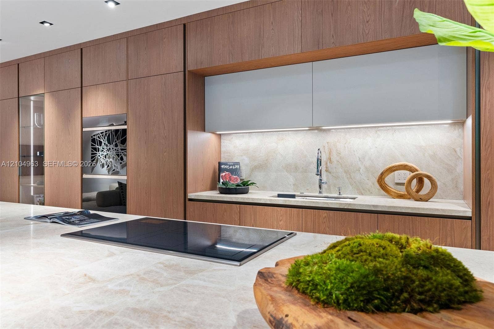 Modern kitchen featuring floor-to-ceiling wood-finish cabinetry, a large stone-surface island with an integrated induction cooktop, light-toned stone backsplash, and recessed ceiling lighting