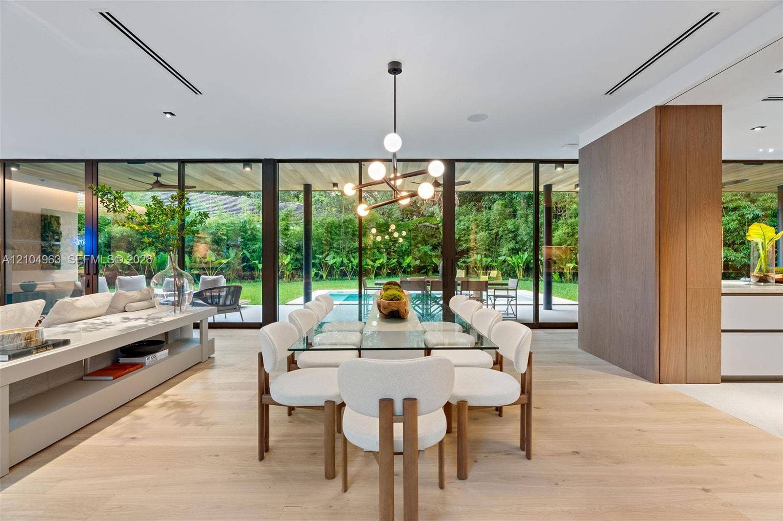 Floor-to-ceiling glass walls, wood-finish flooring, linear recessed lighting, integrated wall cabinetry, and a contemporary multi-arm chandelier