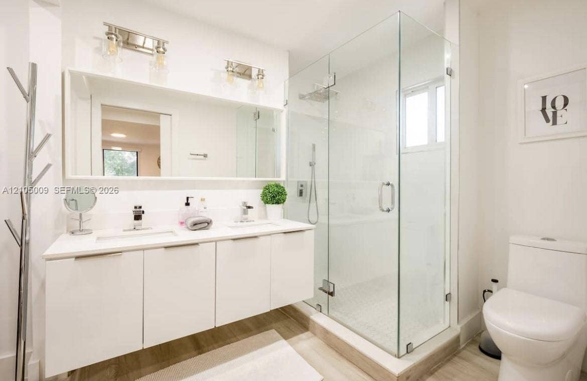 254 NW 51st Street, Miami, Miami, FL 33127 Dual sink vanity with white cabinetry and integrated sinks, large mirrored wall, glass-enclosed shower with tiled walls and floor, and wood-finish flooring