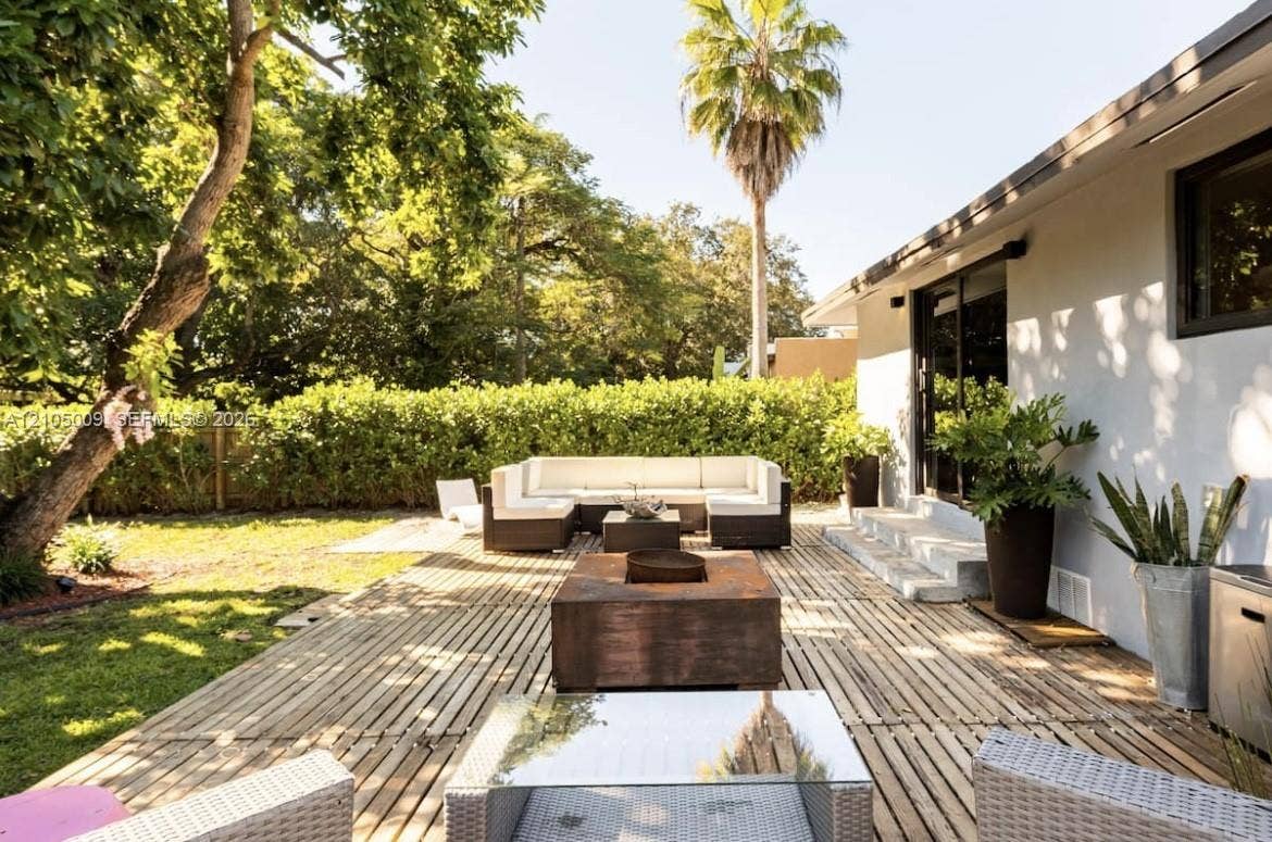 254 NW 51st Street, Miami, Miami, FL 33127 Expansive backyard featuring a wood-slat patio, a manicured lawn, lush perimeter hedging, and a white stucco exterior with dark-framed sliding glass doors