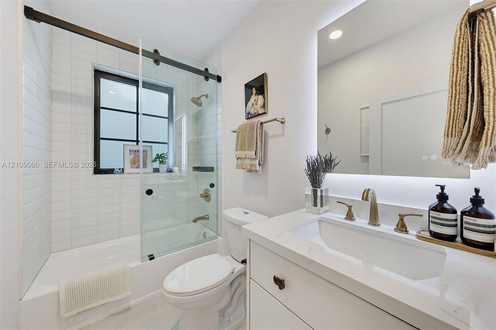 1230 Genoa Street, Coral Gables, Coral Gables, FL 33134 Bathroom featuring a vanity with a white countertop and brass-finish widespread faucet, a backlit mirror, a tub-shower combination with a sliding glass door and black hardware, and subway-style wall tile