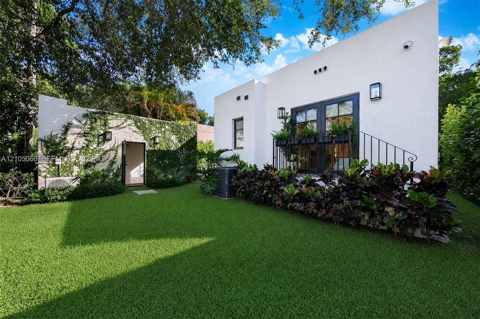 1230 Genoa Street, Coral Gables, Coral Gables, FL 33134 Stucco exterior featuring dual glass-paneled doors, wrought-iron railing, manicured lawn, and lush landscaping