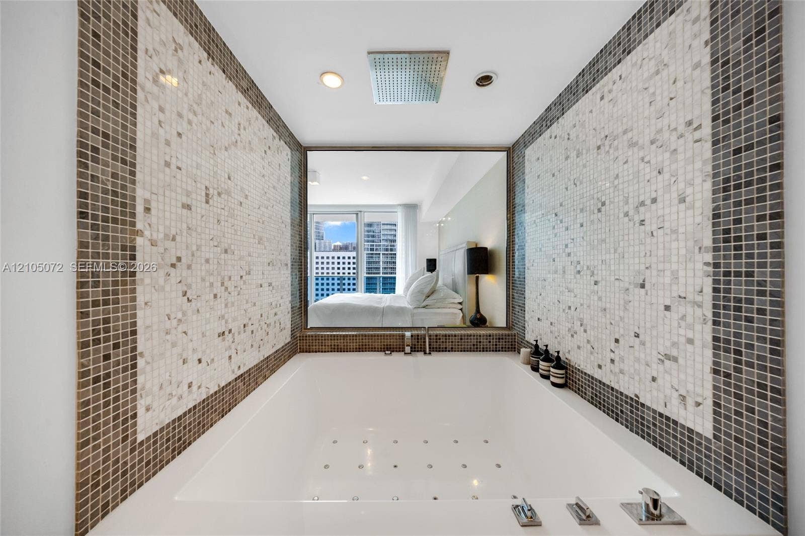 Built-in bathtub framed by mosaic tile and an interior window overlooking the primary bedroom.