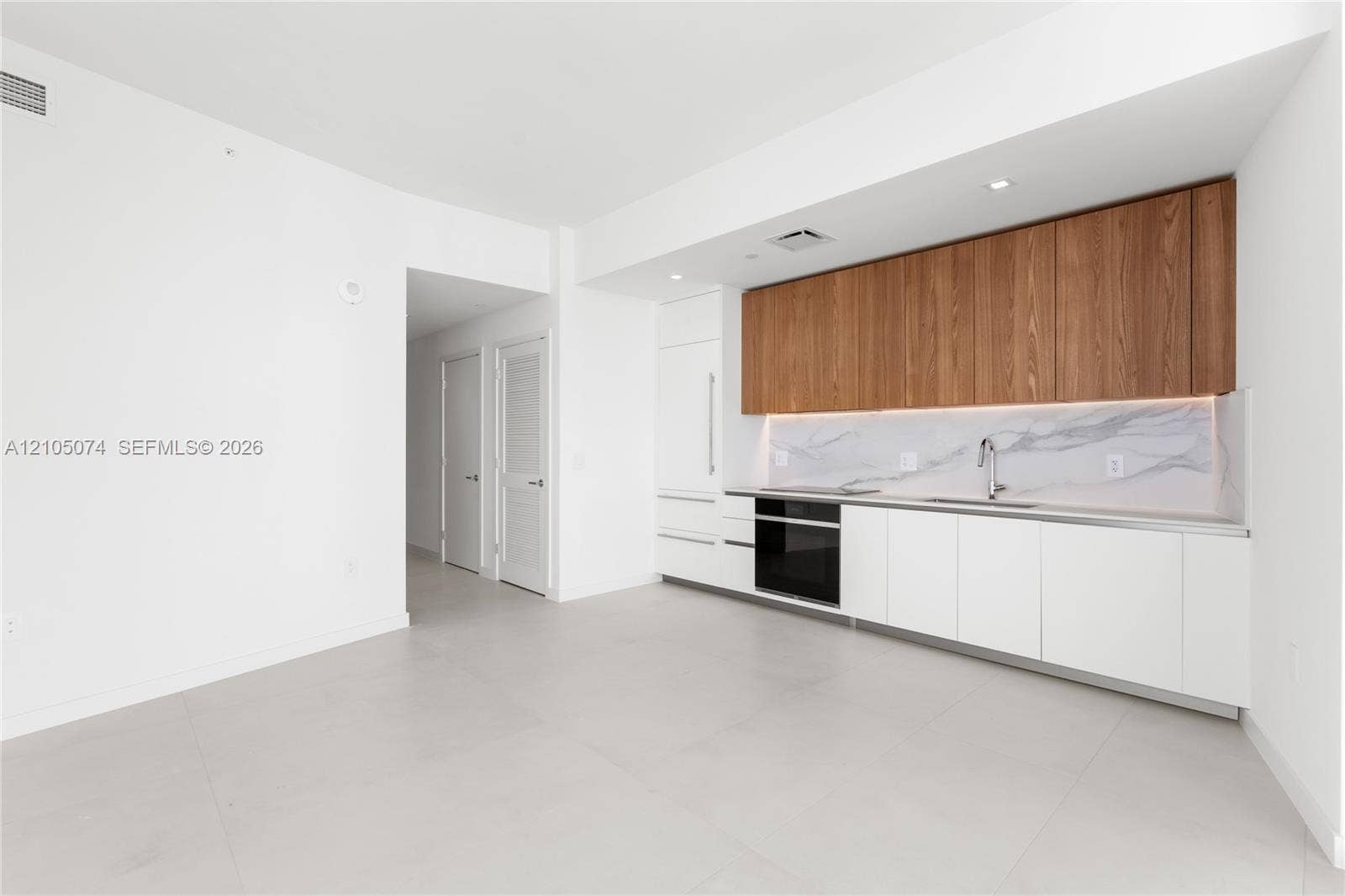Kitchen featuring flat-panel white and wood-finish cabinetry, integrated appliances, stone-patterned backsplash, and light-toned tile flooring