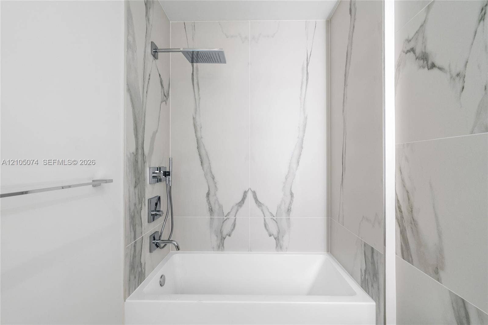 Bathtub featuring large-format veined tile surround, overhead rain shower head, handheld shower wand, and polished chrome fixtures