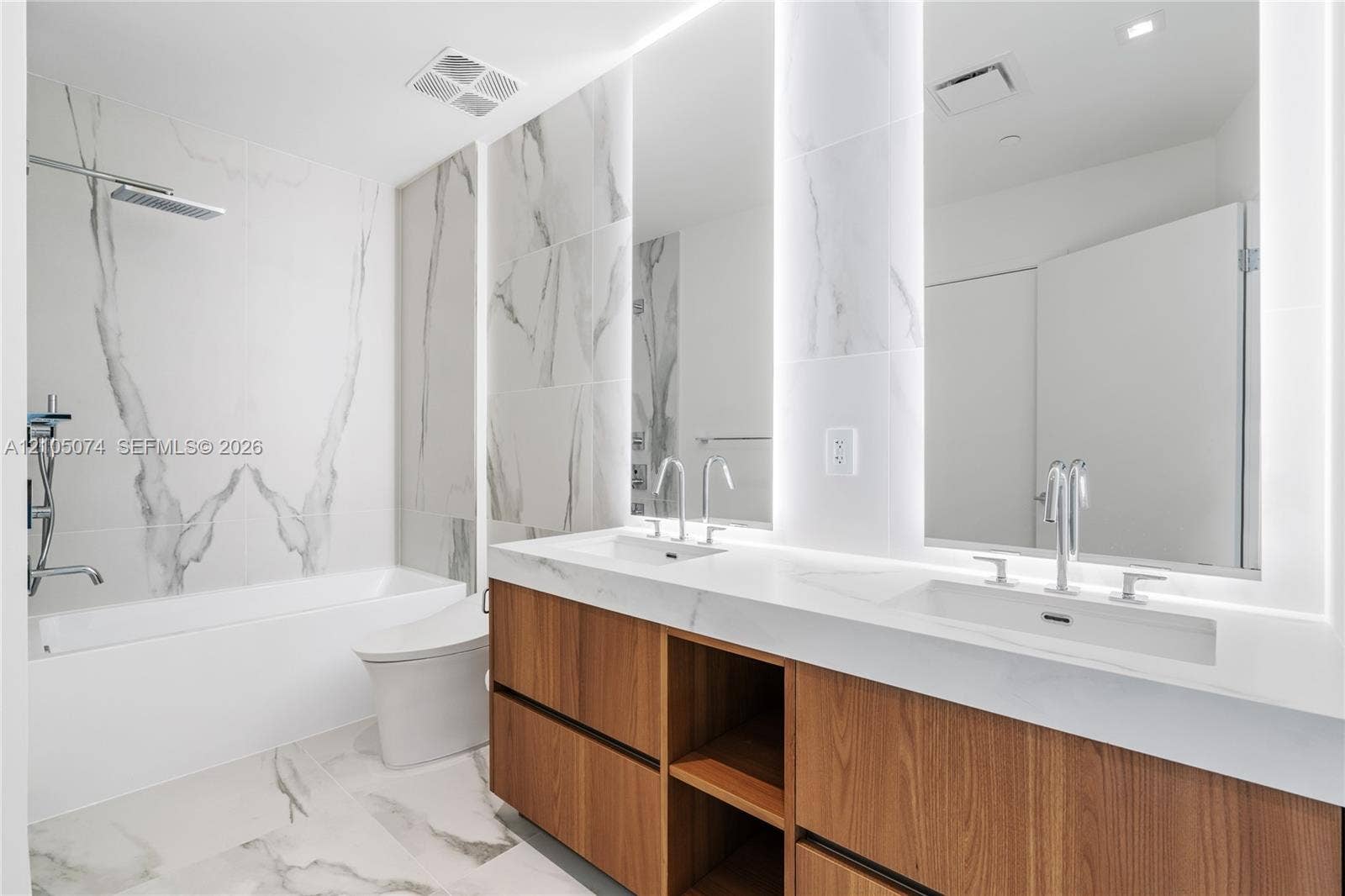Dual-sink vanity with wood-finish cabinetry, quartz countertops, large-format tiled wall surround, undermount tub, and integrated LED mirror lighting