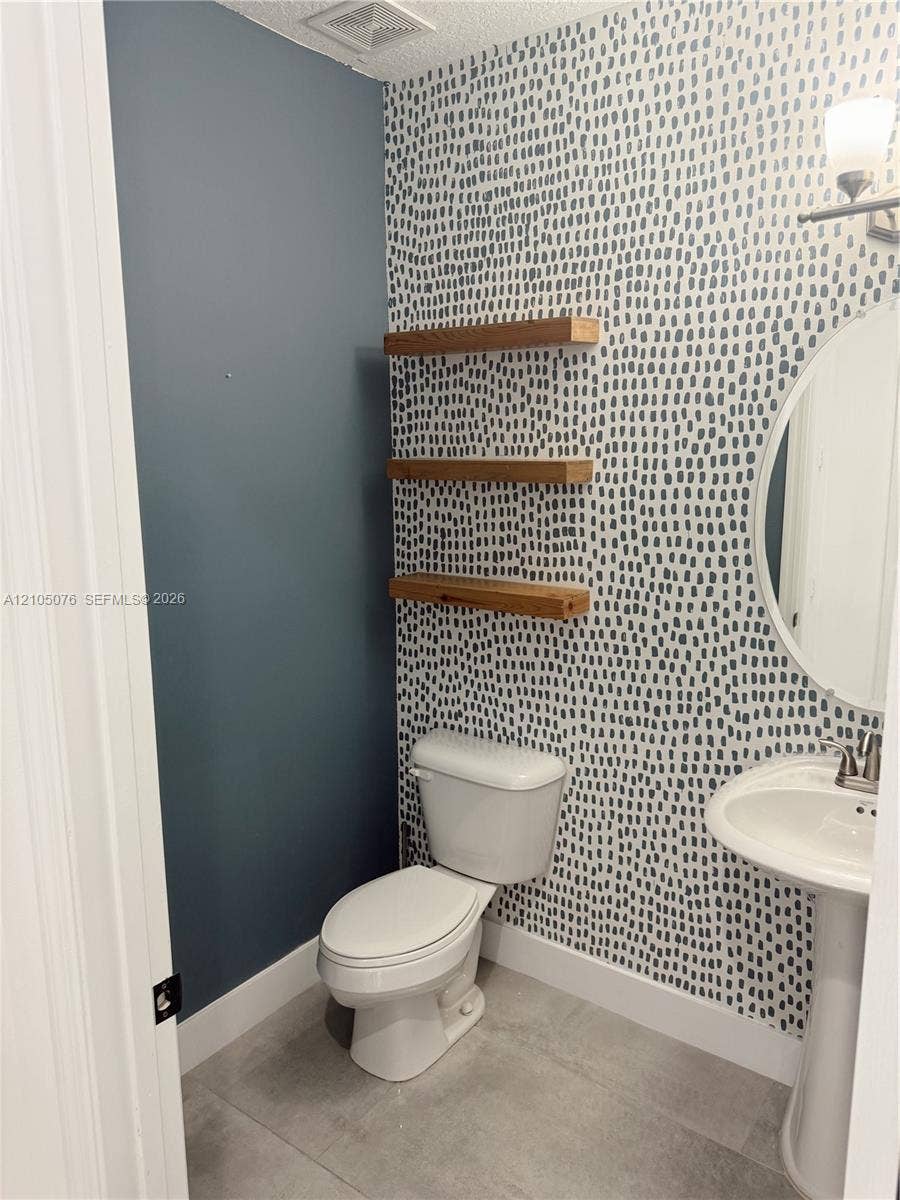 717 NE 191st Street, Miami, FL 33179 Patterned wallpapered accent wall, three built-in wood-finish floating shelves, white pedestal sink with metallic faucet, oval wall-mounted mirror, and tile flooring