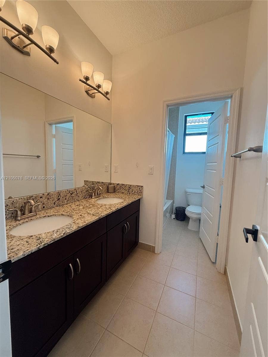 717 NE 191st Street, Miami, FL 33179 Dual-sink vanity with stone-finish countertop, dark wood-finish cabinetry, full-width vanity mirror, tile flooring, and integrated lighting fixtures