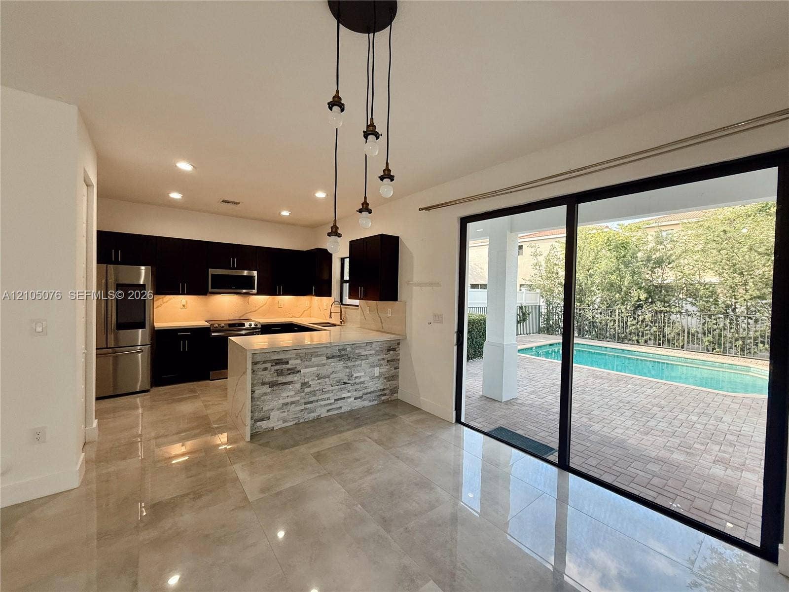 717 NE 191st Street, Miami, FL 33179 Kitchen featuring dark cabinetry, stone-veneer peninsula, recessed lighting, and large-format tile flooring, accompanied by expansive sliding glass doors opening to a paved patio and swimming pool