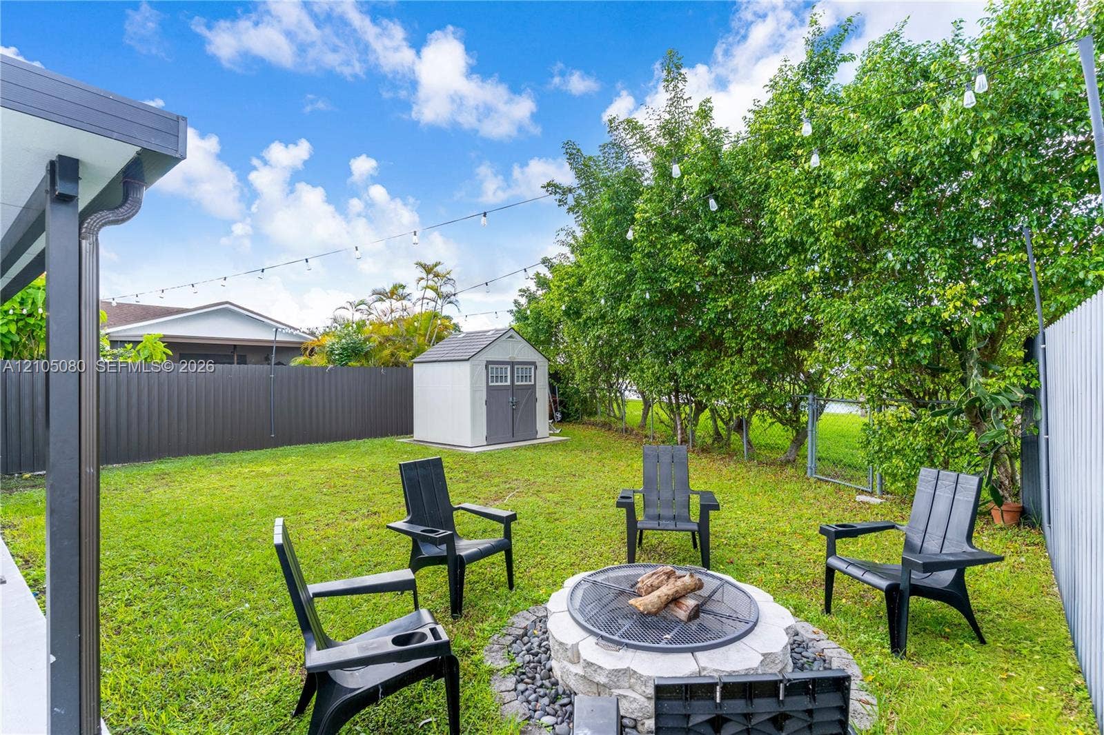 9755 SW 148th Court, Miami, FL 33196 Backyard featuring a stone-encased fire pit, detached storage shed, privacy fencing, and overhead string lighting