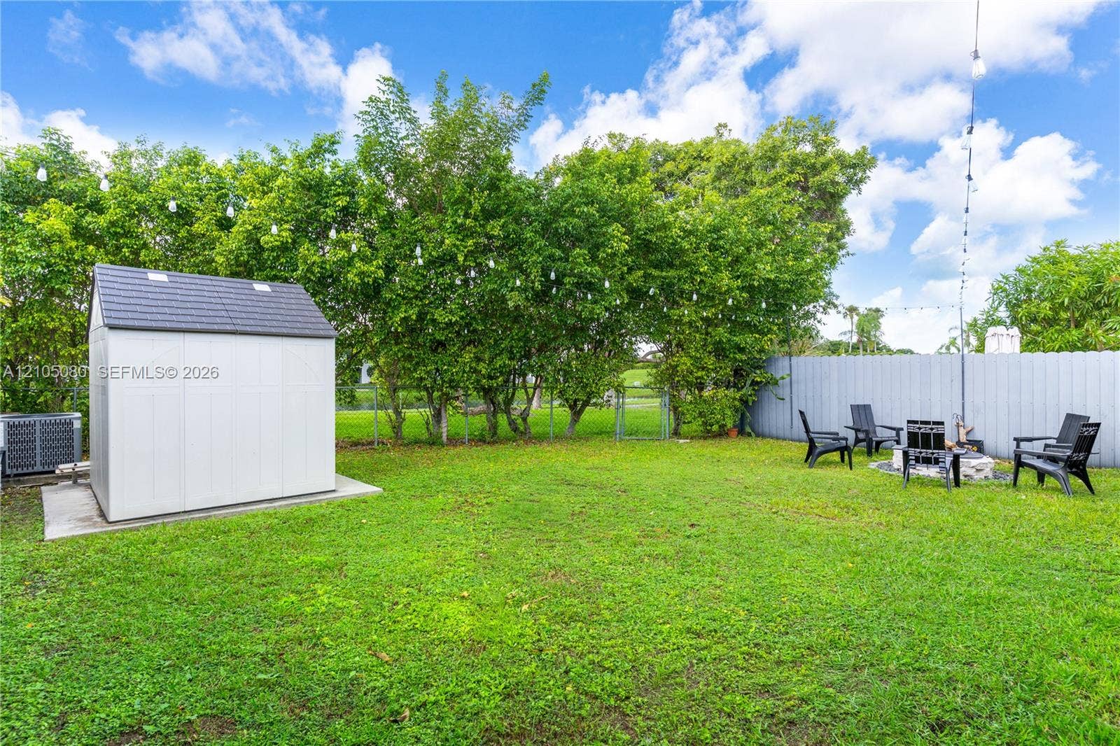 9755 SW 148th Court, Miami, FL 33196 Fenced backyard featuring a manicured lawn, detached storage structure on a concrete pad, mature perimeter trees, and string lighting