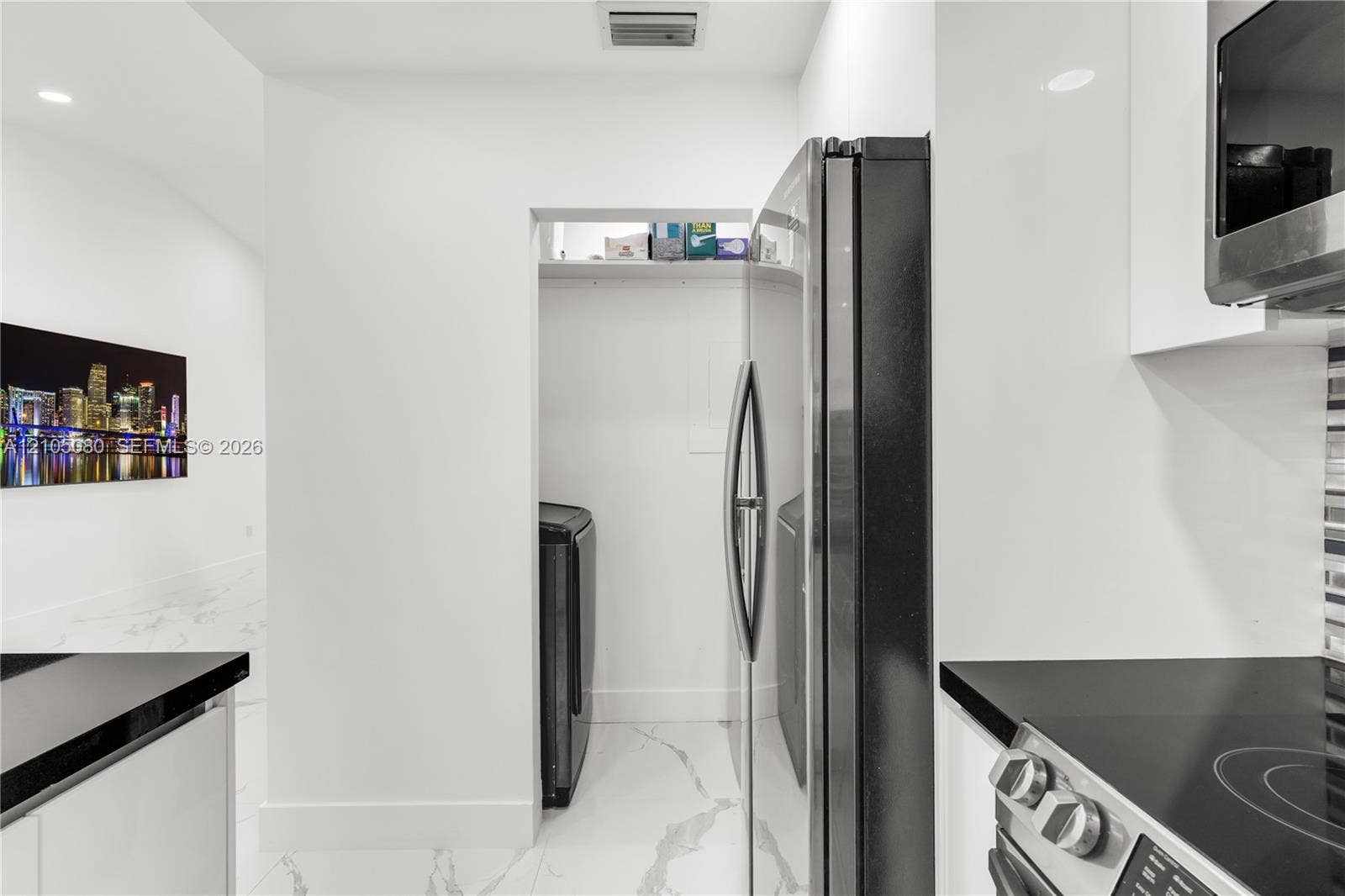 9755 SW 148th Court, Miami, FL 33196 Kitchen featuring high-gloss white cabinetry, stone-finish tile flooring, a dark-toned integrated refrigerator, and a recessed laundry alcove with overhead shelving