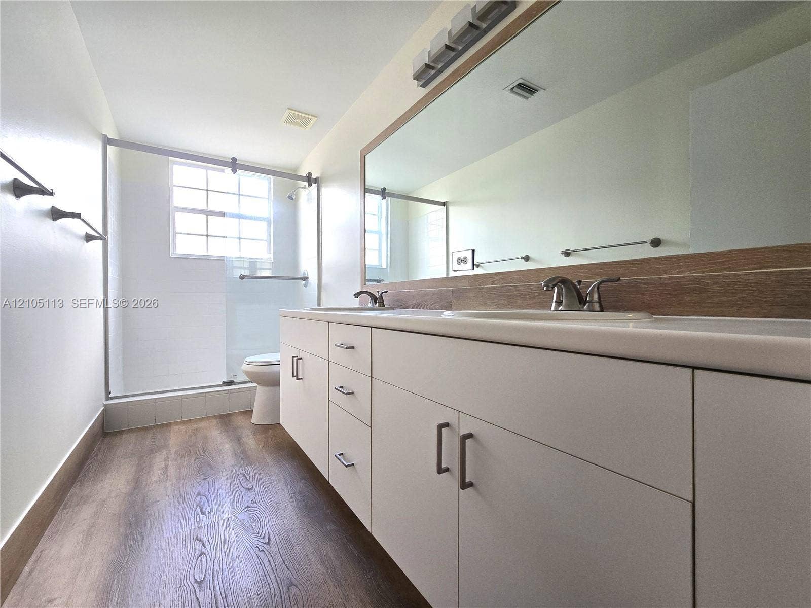 2020 Pompeii Court, Weston, Weston, FL 33327 Dual vanity with integrated sinks and chrome fixtures, white cabinetry with dark pulls, full-width mirror with wood-finish trim, glass-enclosed shower, and wood-finish flooring