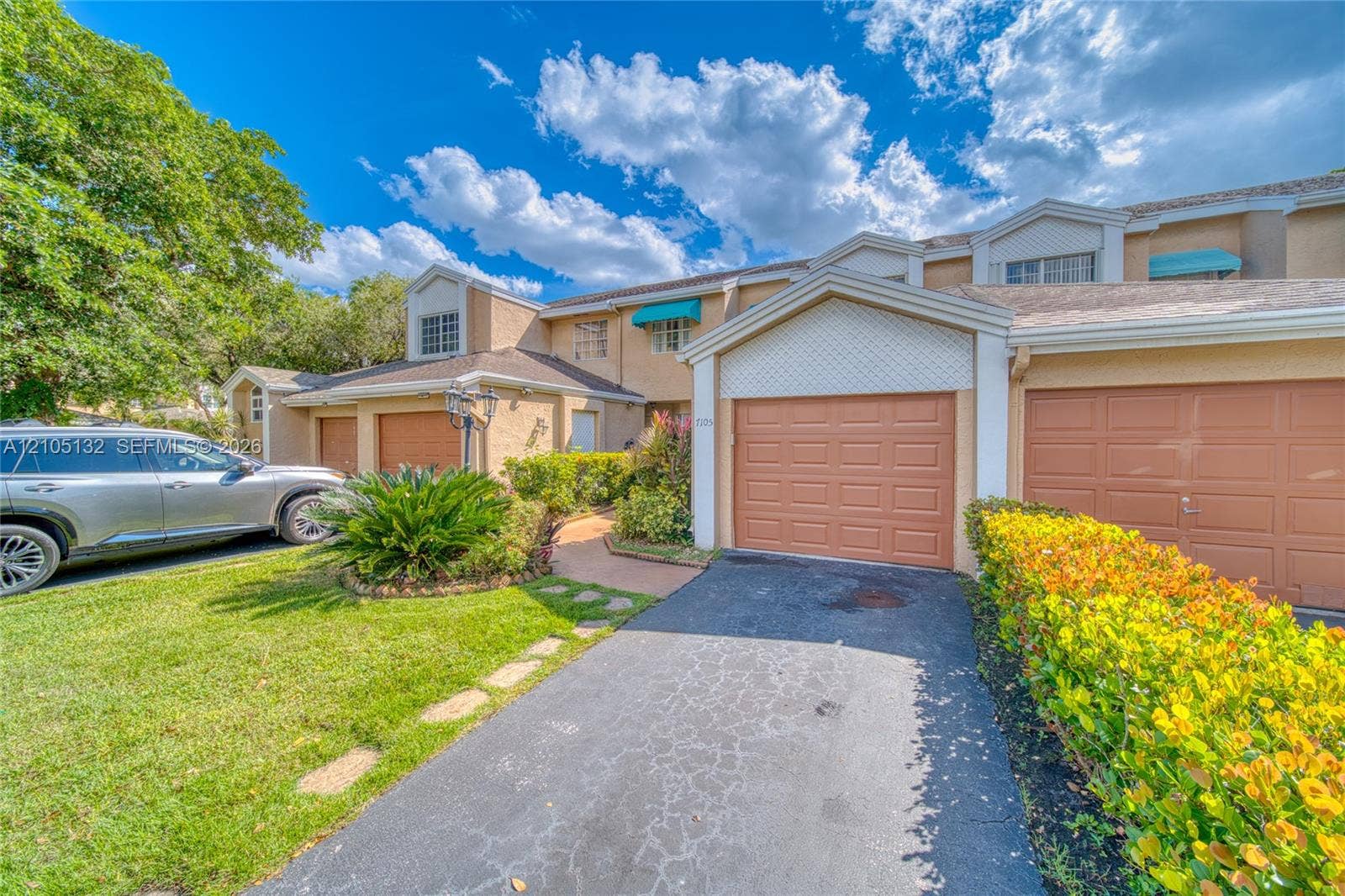 Tiled roofline with dormer windows, stucco exterior, multiple garage doors, manicured shrubbery, and paved driveway