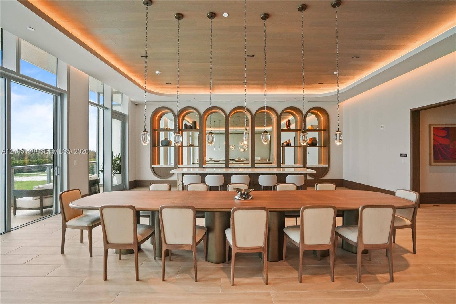 2411 Laguna Circle, #1505, North Miami, FL 33181 Open-concept dining area featuring a long wood dining table, recessed ceiling lighting, a built-in bar with arched shelving and mirrored accents, and full-height glass doors