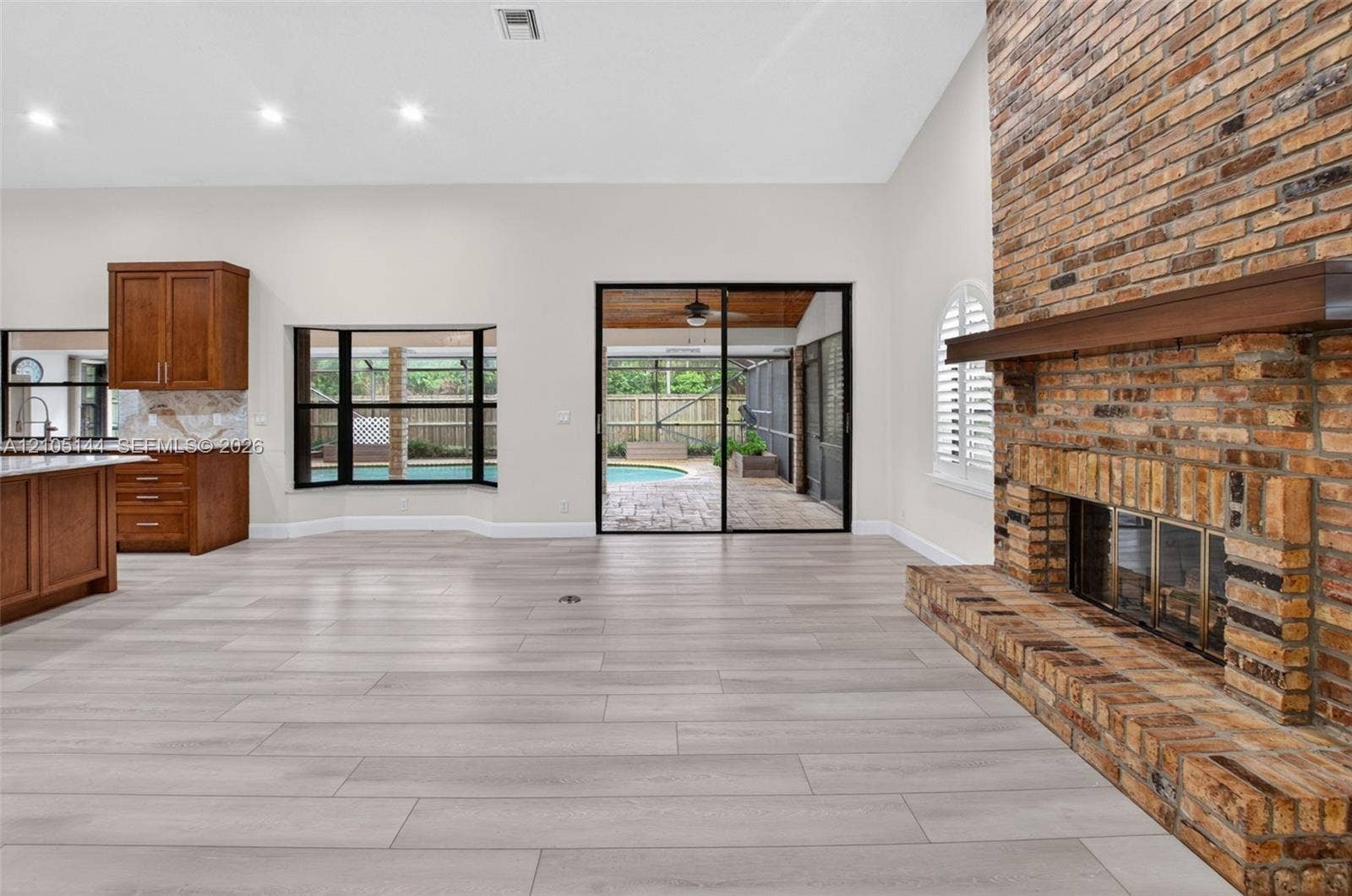 Vaulted ceiling living area featuring a full-height exposed brick fireplace, wood-finish flooring, recessed lighting, and sliding glass doors leading to a screened-in lanai