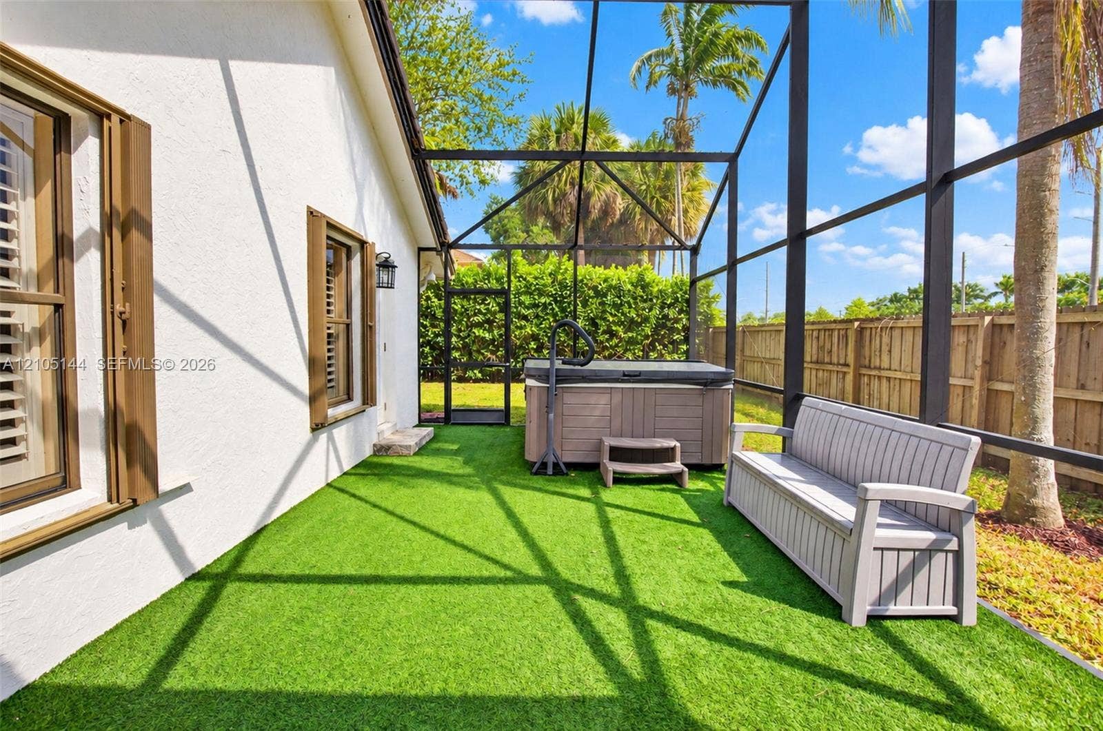 Screened lanai featuring synthetic grass flooring, a built-in hot tub, and a wood-tone fence enclosure