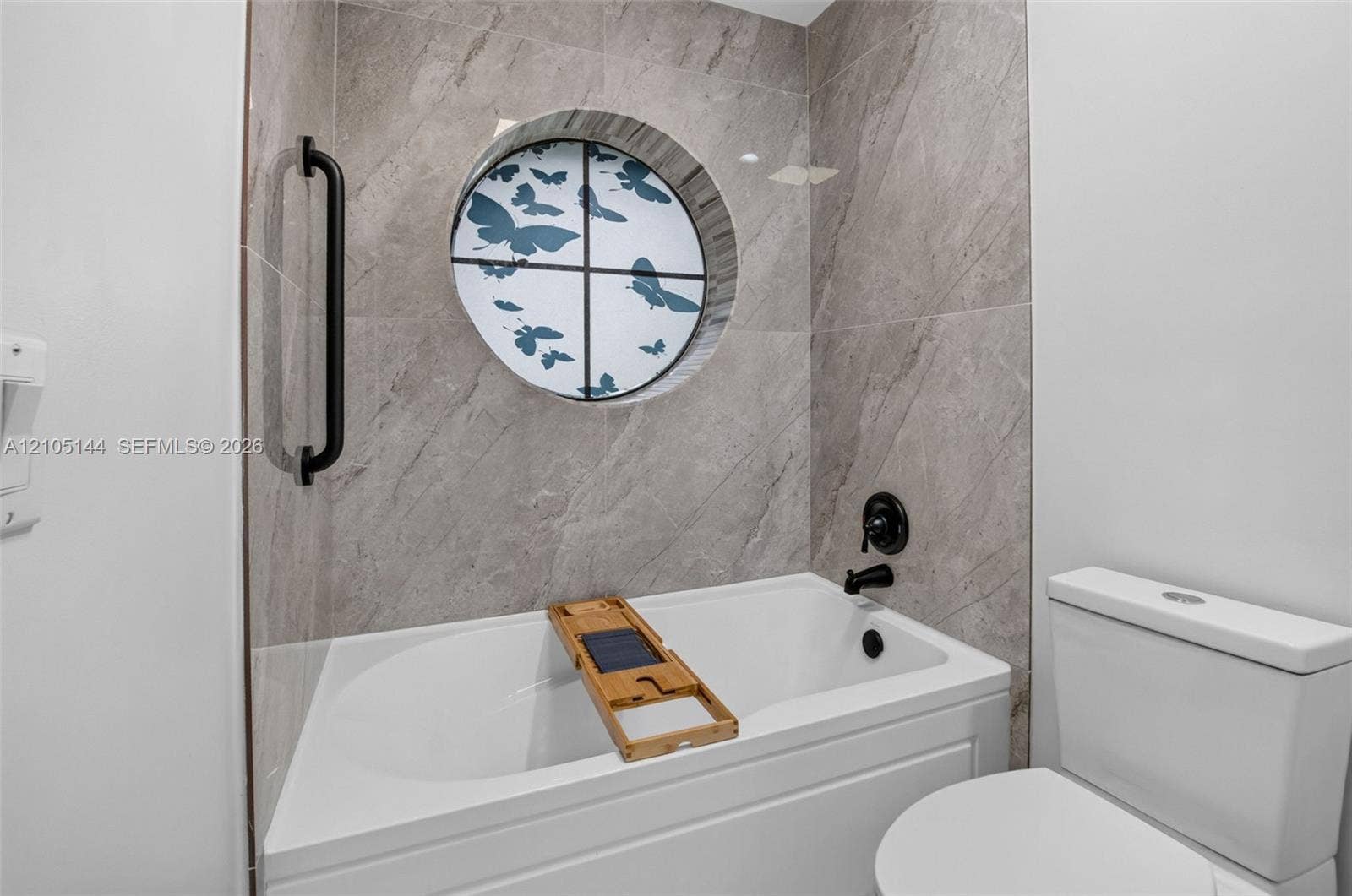 Bathtub featuring large-format grey tile surround, circular accent window, matte black faucet fixtures, and wall-mounted grab bar