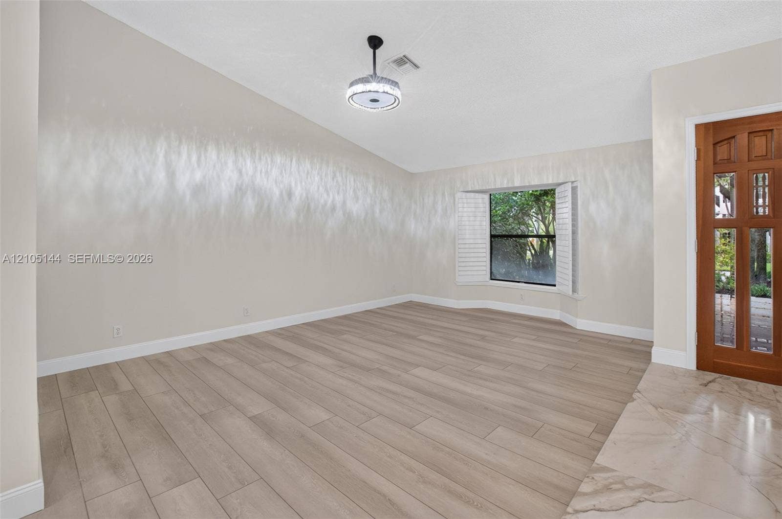 Vaulted ceilings, wood-finish flooring, recessed bay window with plantation shutters, integrated circular flush-mount lighting, and decorative wood-paneled entry door