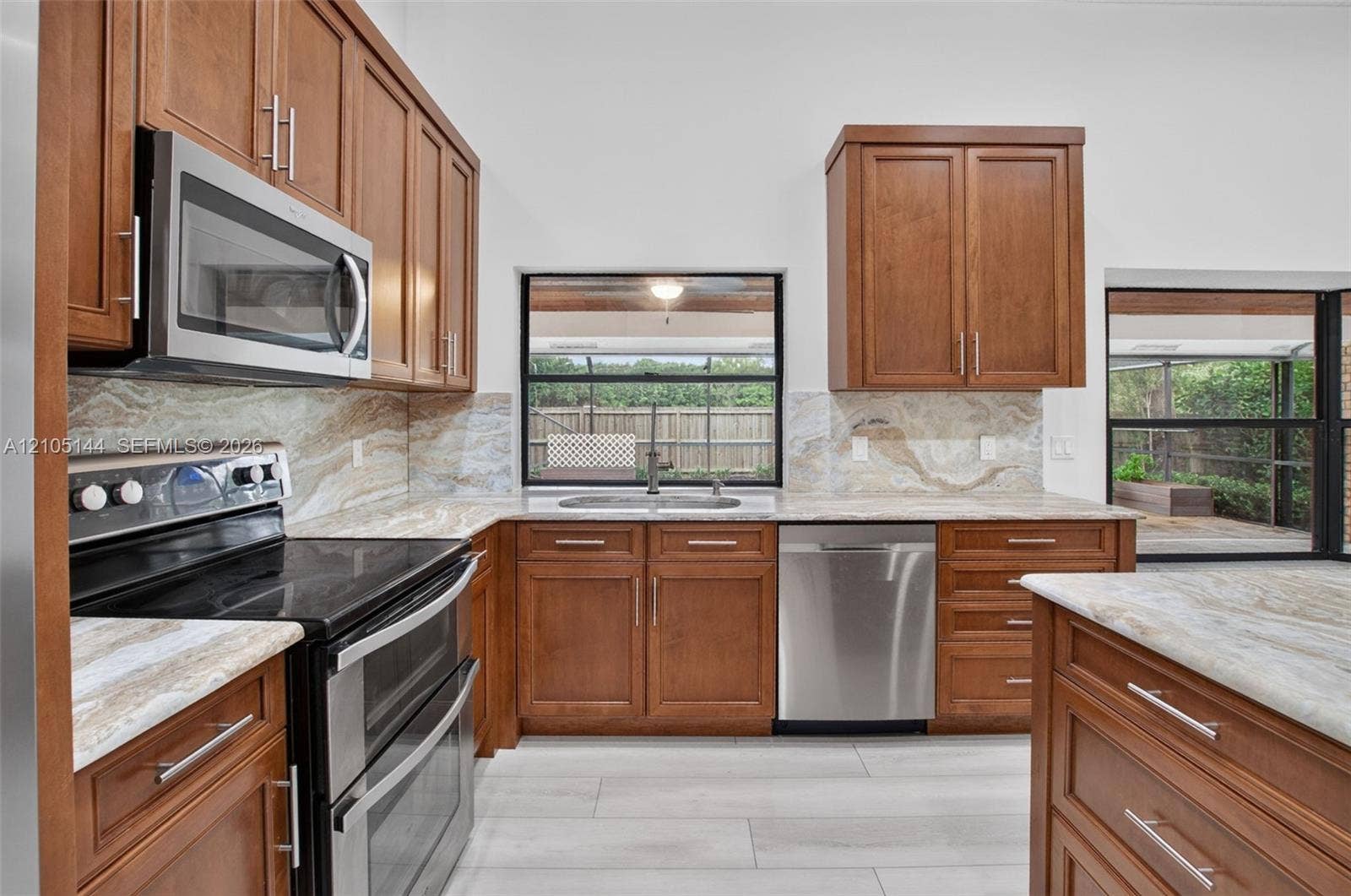 Kitchen featuring medium-tone wood cabinetry, stone-finish countertops with matching full-height backsplash, stainless steel appliances, and large-format tile flooring