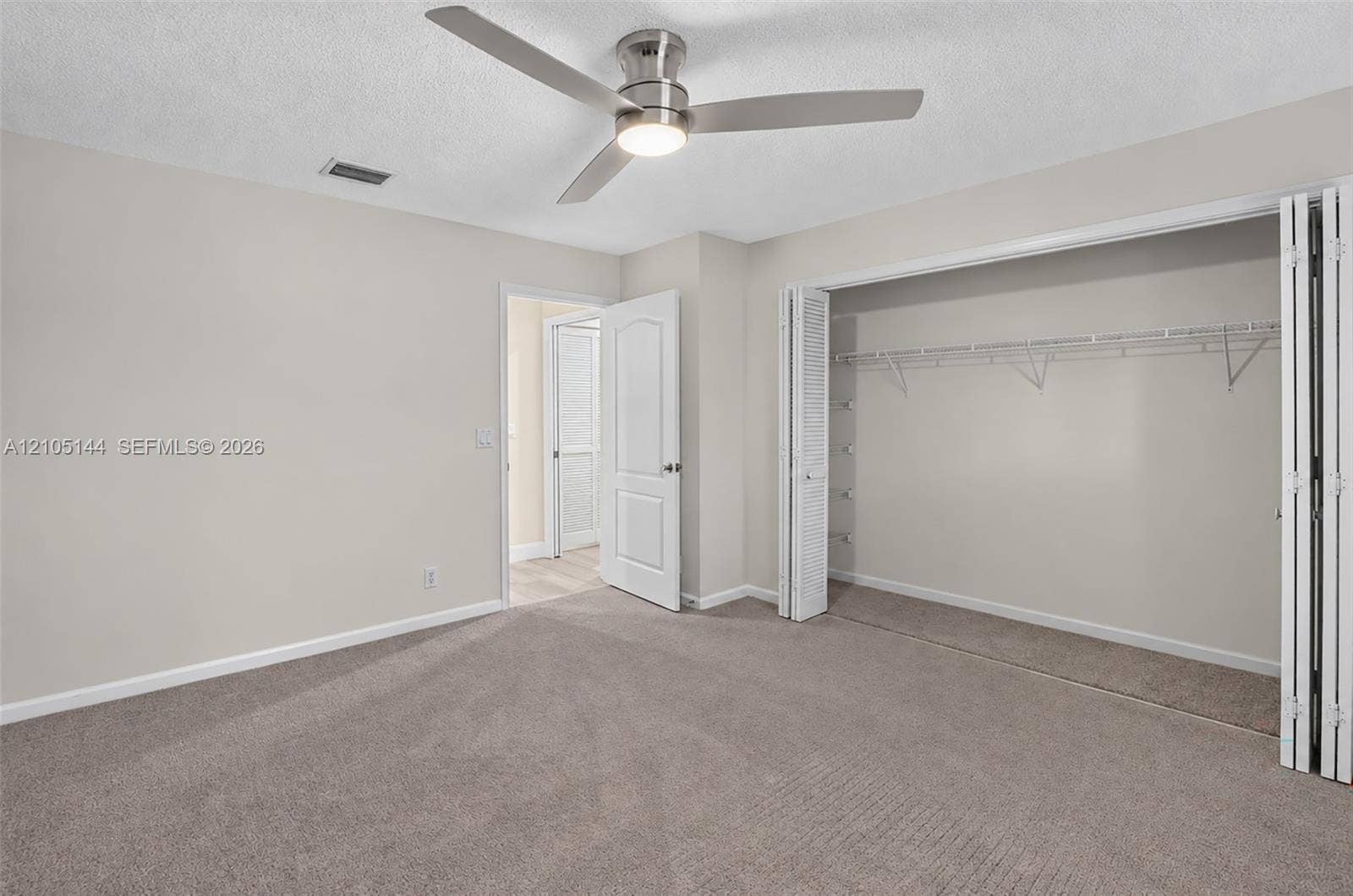 (FAN VIRTUALLY STAGED AS SIMILAR PIECE WILL BE INSTALLED) Neutral-toned interior space featuring neutral wall-to-wall carpeting, a satin-nickel finish ceiling fan with integrated lighting, and a closet with bifold doors and built-in shelving