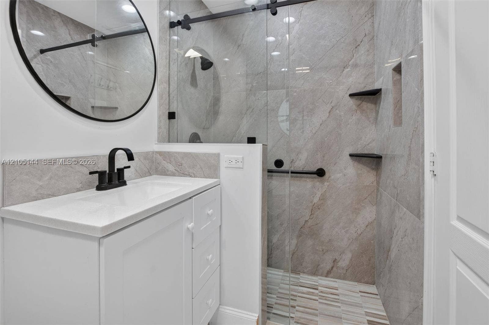 White vanity cabinet with an integrated rectangular sink and matte black faucet, paired with a frameless glass shower enclosure featuring large-format stone-tile walls, built-in shelving, and matte black hardware