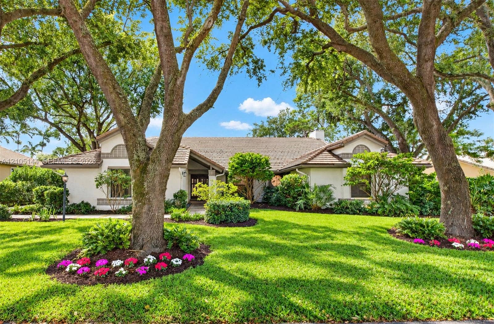 Stucco exterior featuring arched architectural details, a tiled roof, mature canopy trees, and landscaped garden beds with seasonal plantings