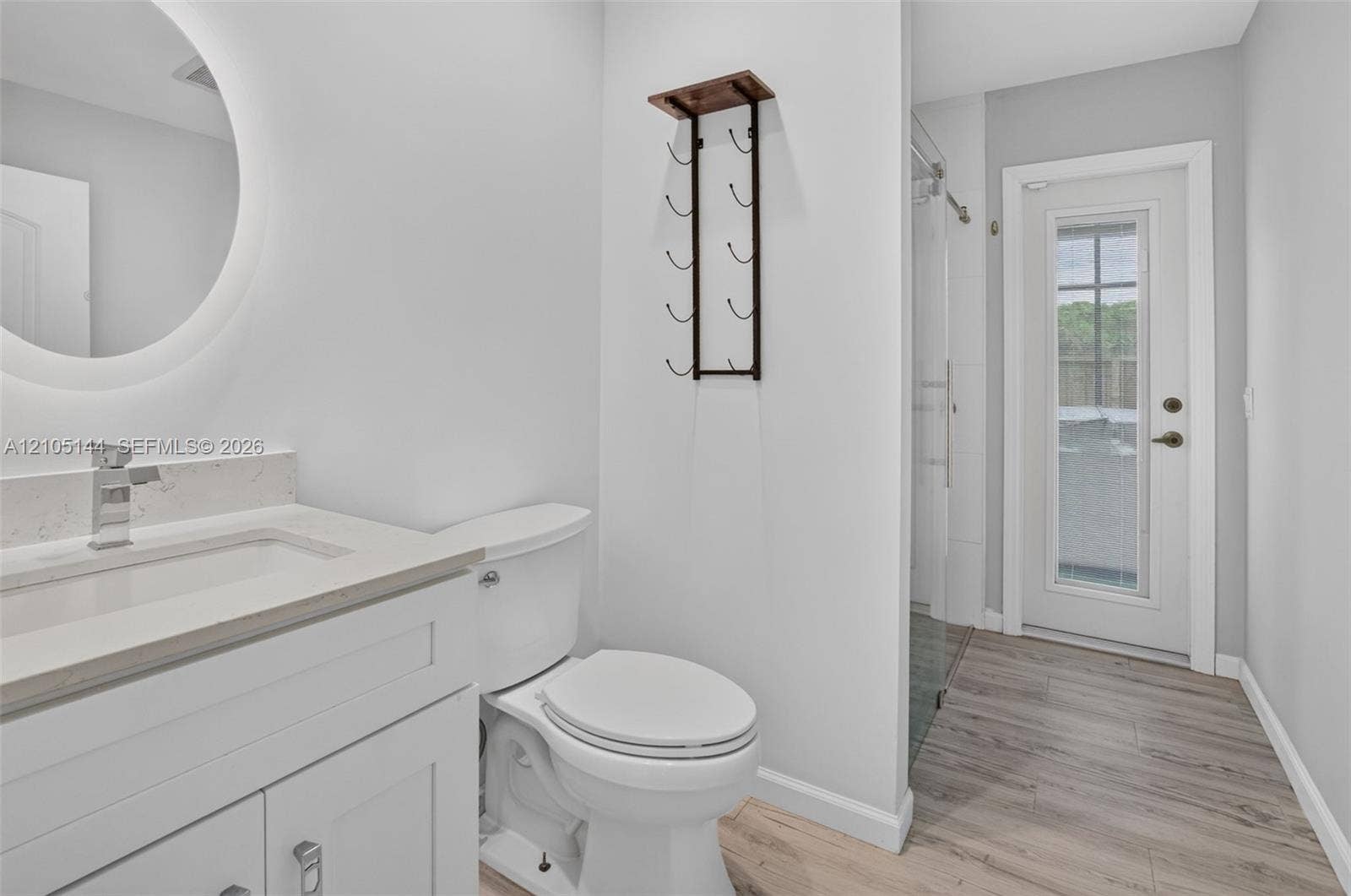 Bathroom featuring a white vanity with a light-toned stone countertop, integrated sink with a chrome faucet, circular LED-backlit mirror, and wood-finish flooring
