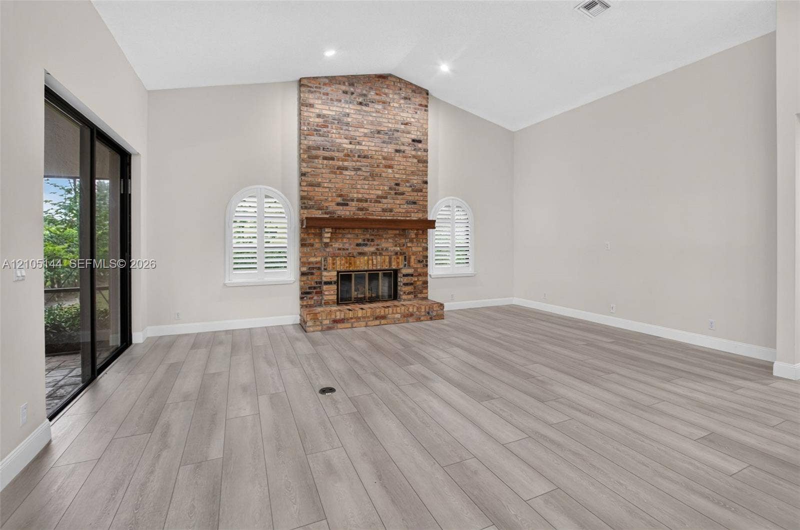 Vaulted ceiling, floor-to-ceiling brick fireplace with wood mantel, wood-finish flooring, arched windows with plantation shutters, and sliding glass door