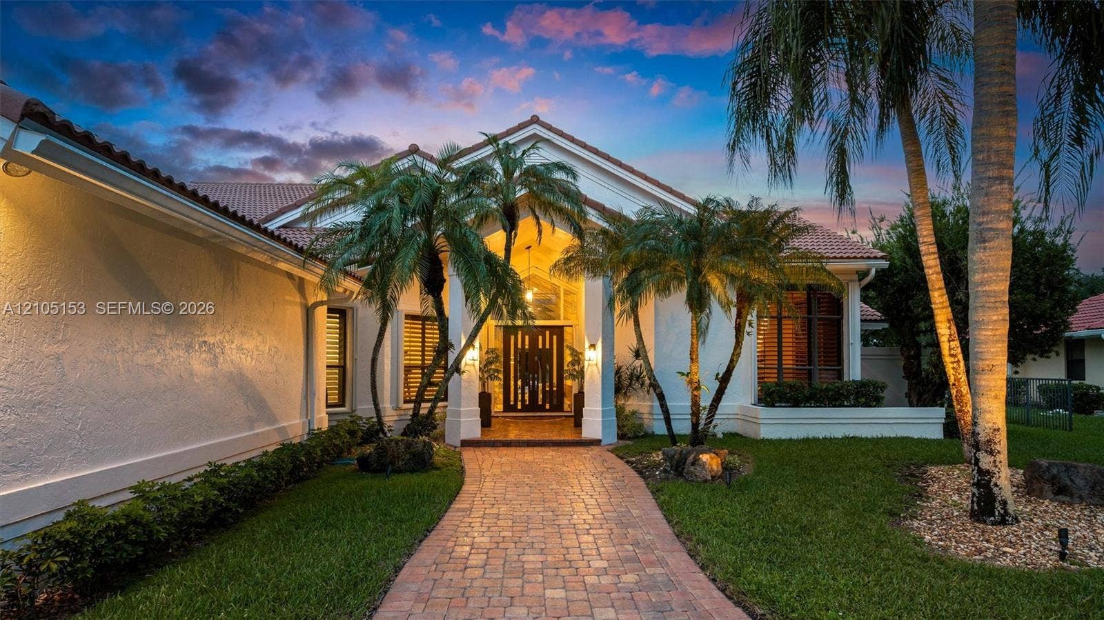 Paver walkway leading to a grand entry, surrounded by mature palm trees, a well-maintained lawn, and stucco exterior with a tile roof