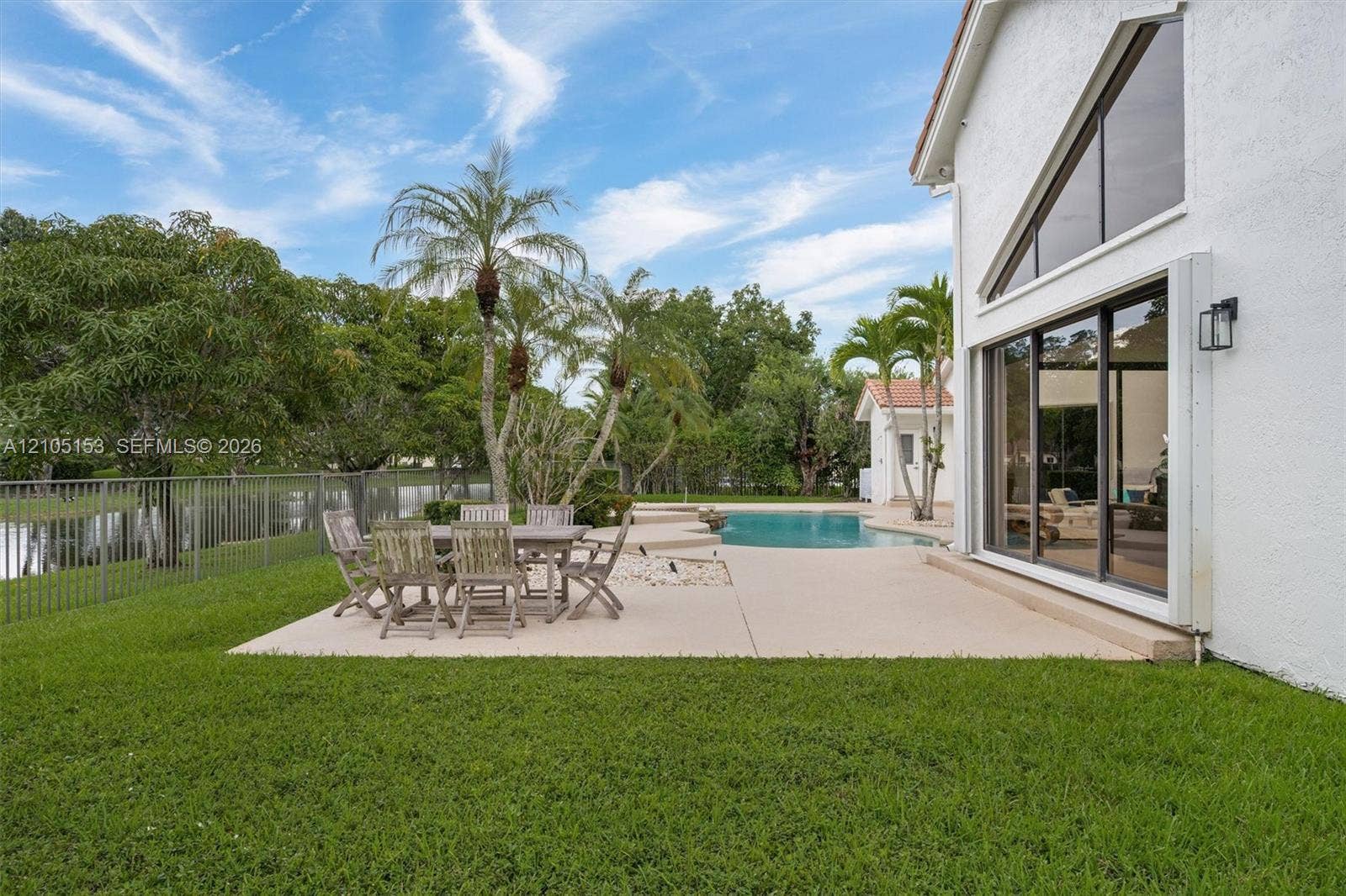 Private swimming pool with concrete decking, surrounding green space, mature palm trees, and a perimeter fence
