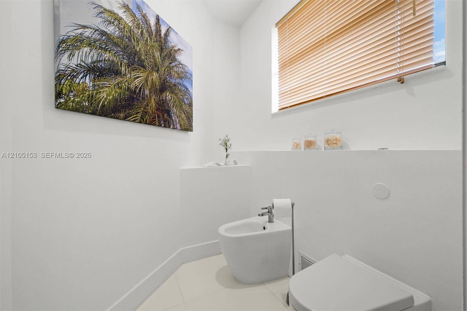 White bidet and toilet, neutral tile flooring, and a window with wood-finish blinds
