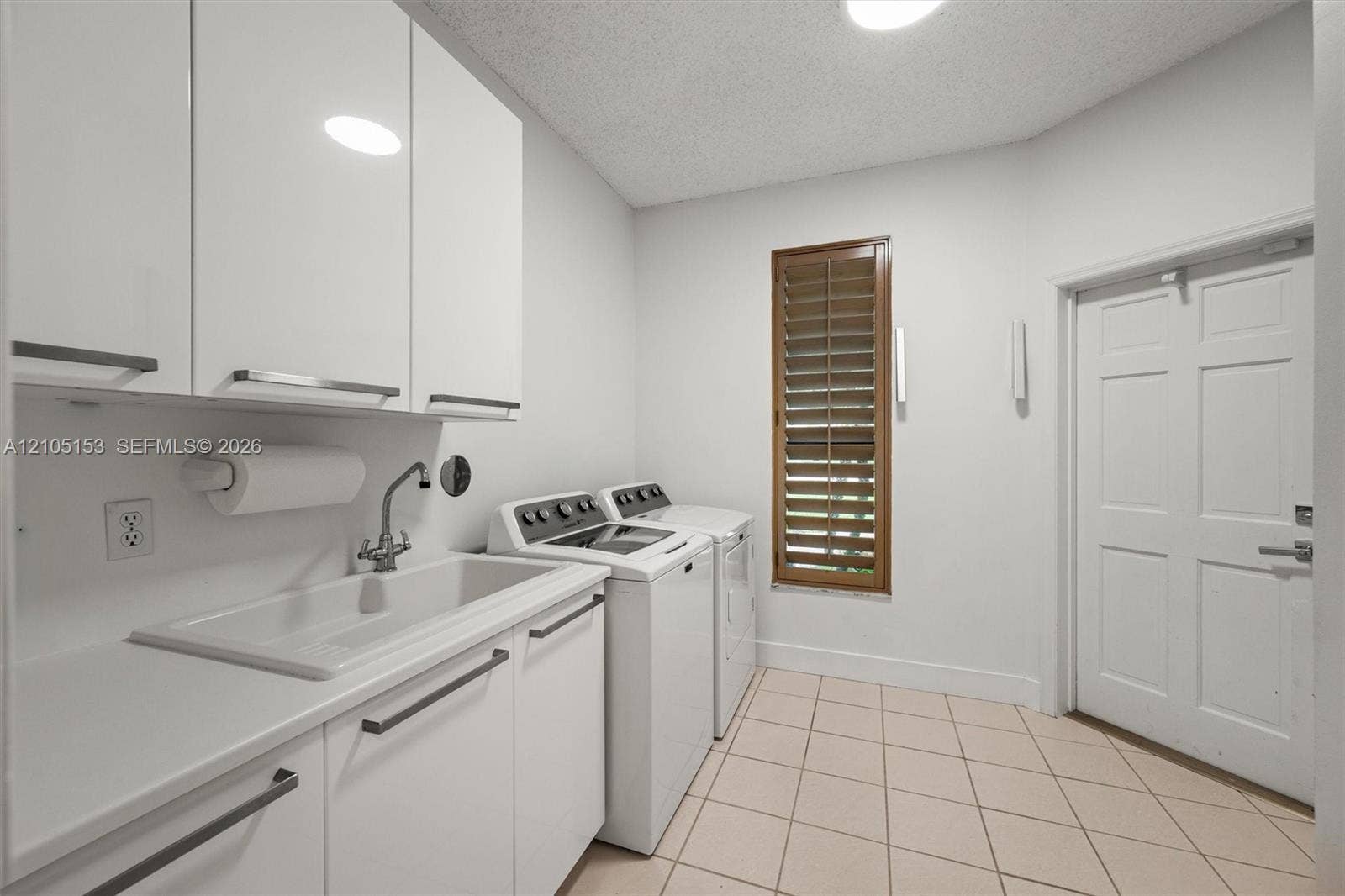 White cabinetry with linear hardware, integrated utility sink, wood-finish louvered window treatments, white paneled door, and tiled flooring