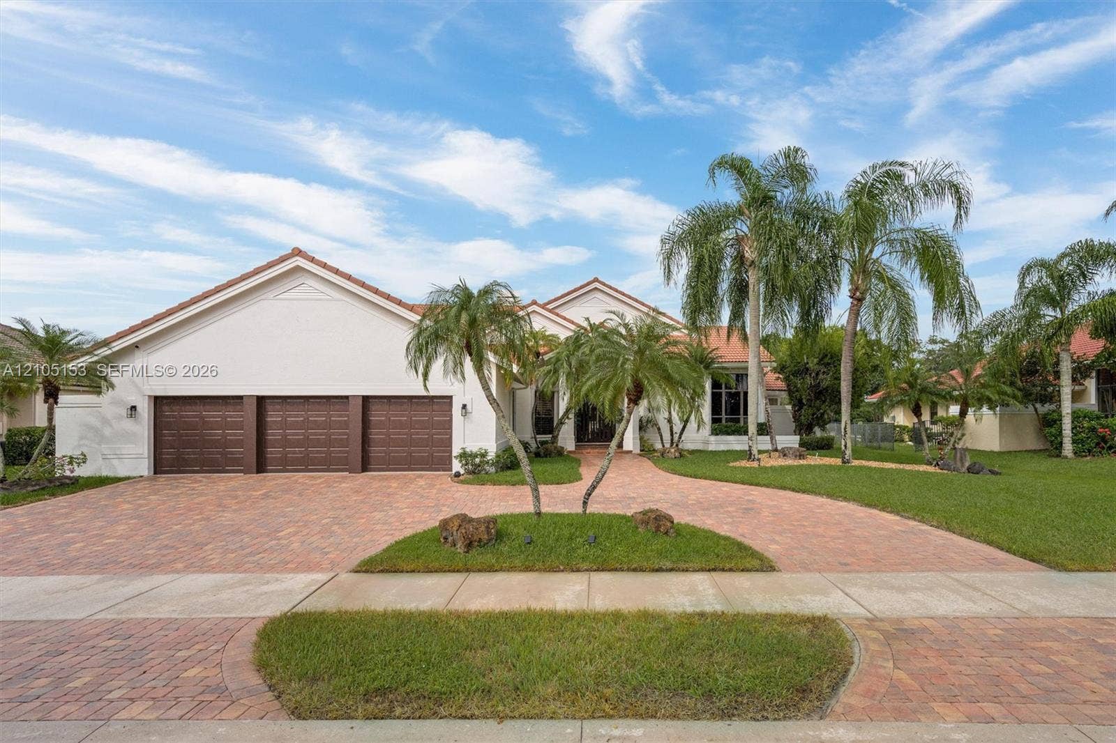 Stucco exterior with a tile roof, three-car garage, brick paver driveway, mature palm trees, and a landscaped lawn