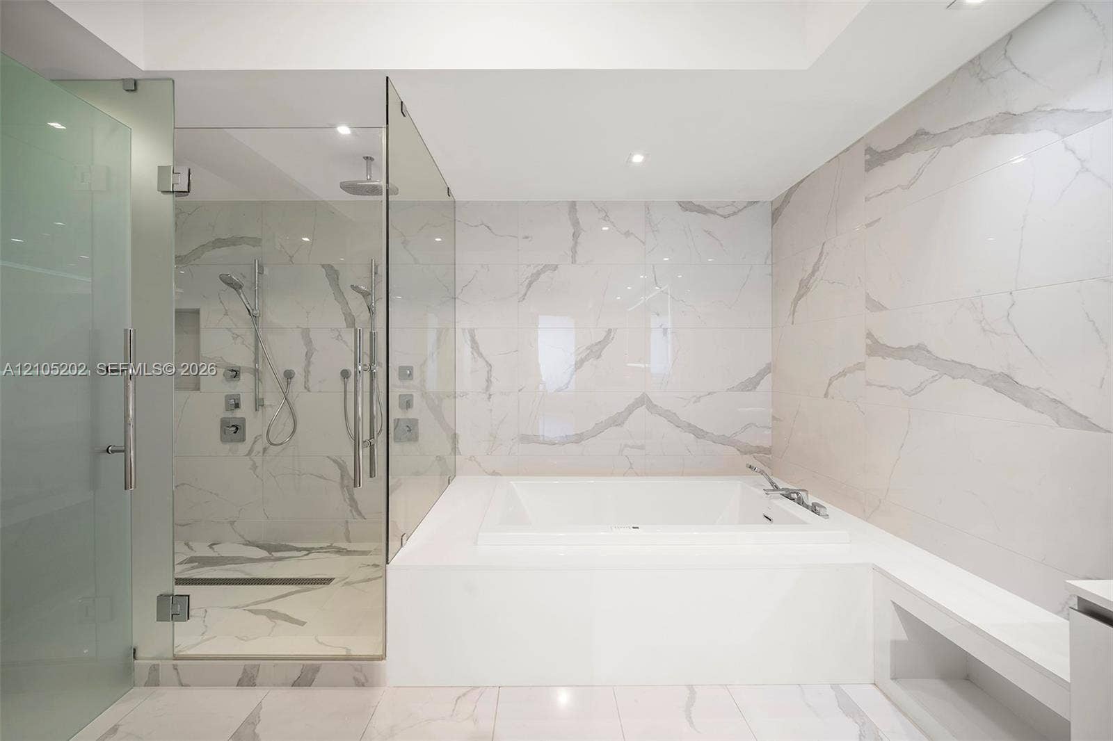 Enclosed glass shower featuring dual shower heads and a built-in recessed niche, complemented by a deep soaking tub surrounded by full-height stone-finish wall tiling and matching floor tile
