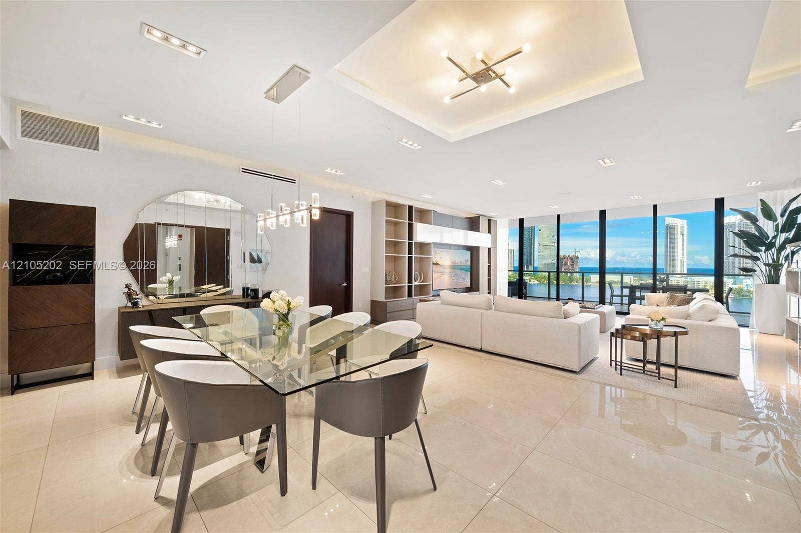 Open-concept interior featuring large-format tile flooring, tray ceilings with recessed lighting, custom built-in cabinetry, and floor-to-ceiling windows with water-centric scenery