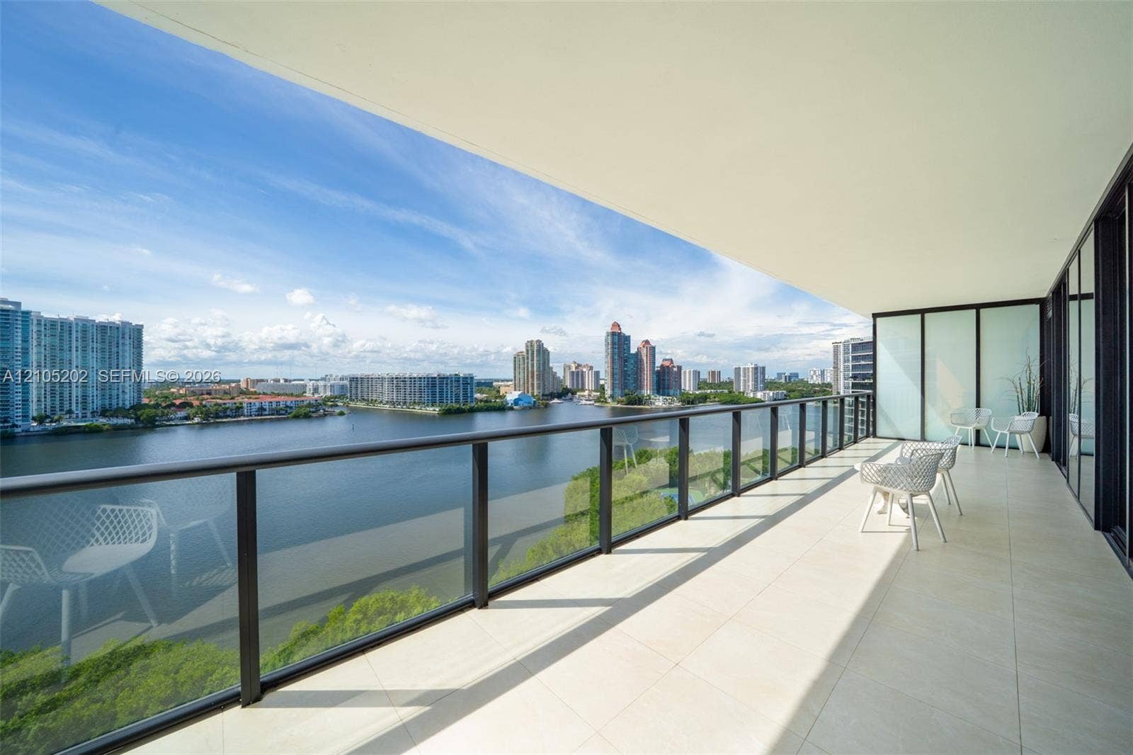 Expansive covered balcony featuring large-format tile flooring and a glass-panel railing with dark metal framing, showcasing a water and city skyline backdrop