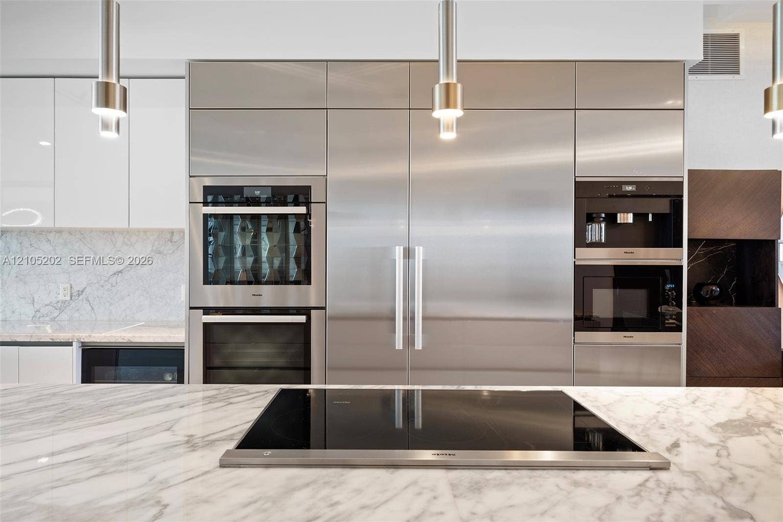 Modern kitchen featuring integrated stainless steel appliances, stone slab countertops, a built-in induction cooktop, high-gloss cabinetry, and pendant lighting