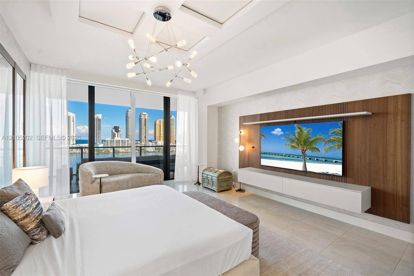 Floor-to-ceiling glass windows, custom wood-paneled accent wall with integrated floating media console, modern multi-arm chandelier, tray ceiling detailing, and large-format tile flooring