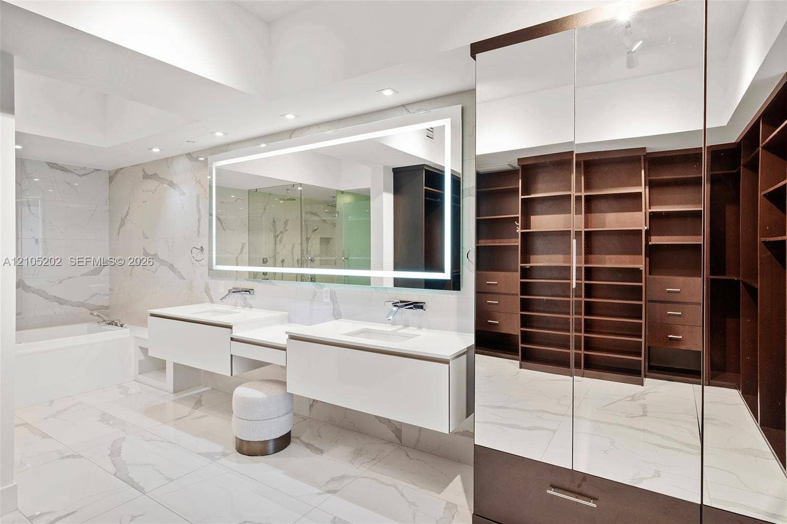 Floating dual vanity with integrated sinks, large LED-backlit mirror, floor-to-ceiling tile, built-in wardrobe cabinetry with mirrored doors, and deep soaking tub