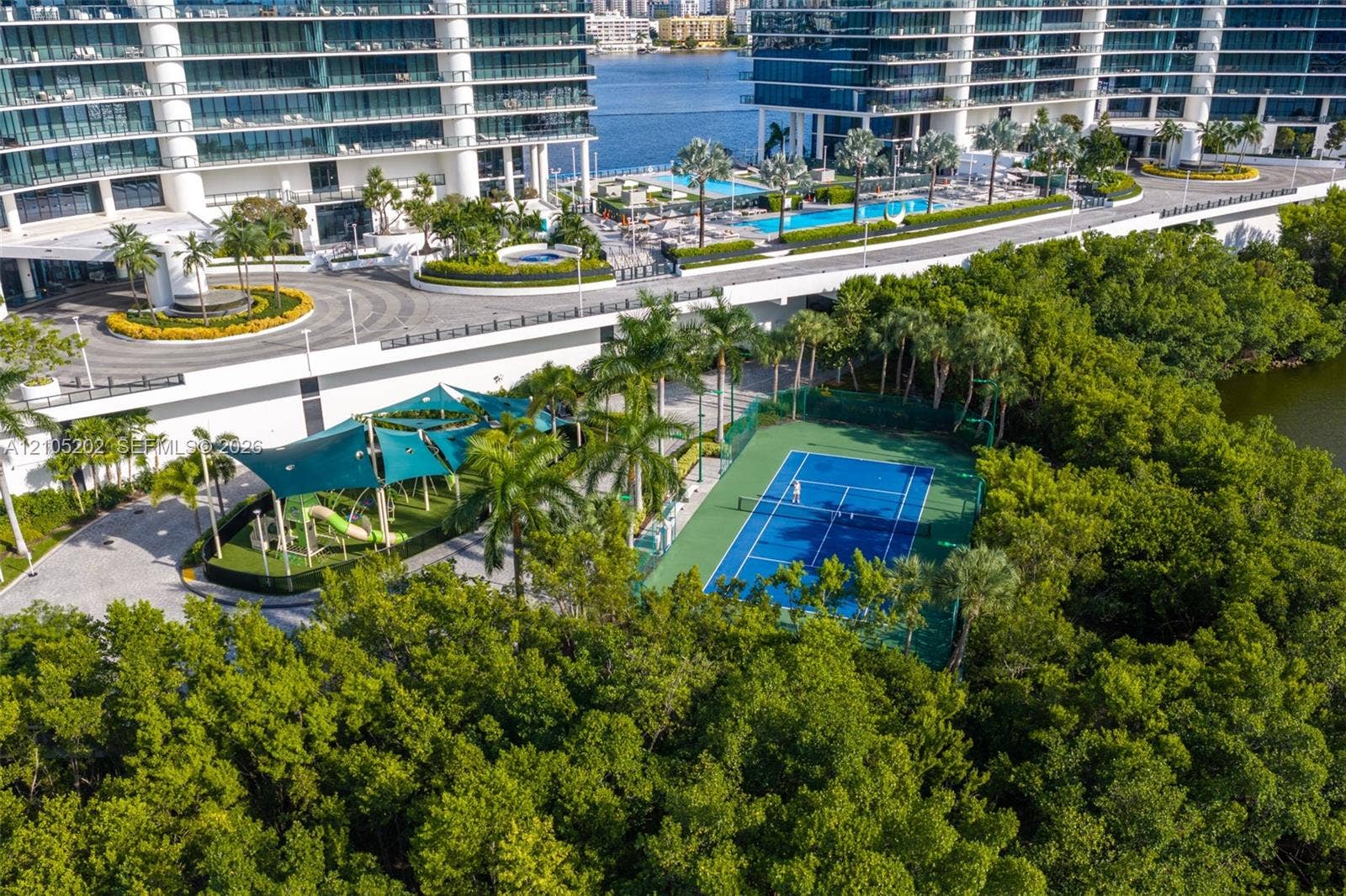 Aerial view of residential grounds featuring a multi-use hard-surface tennis court, a shade-covered recreation structure, mature palm landscaping, and paved pedestrian walkways