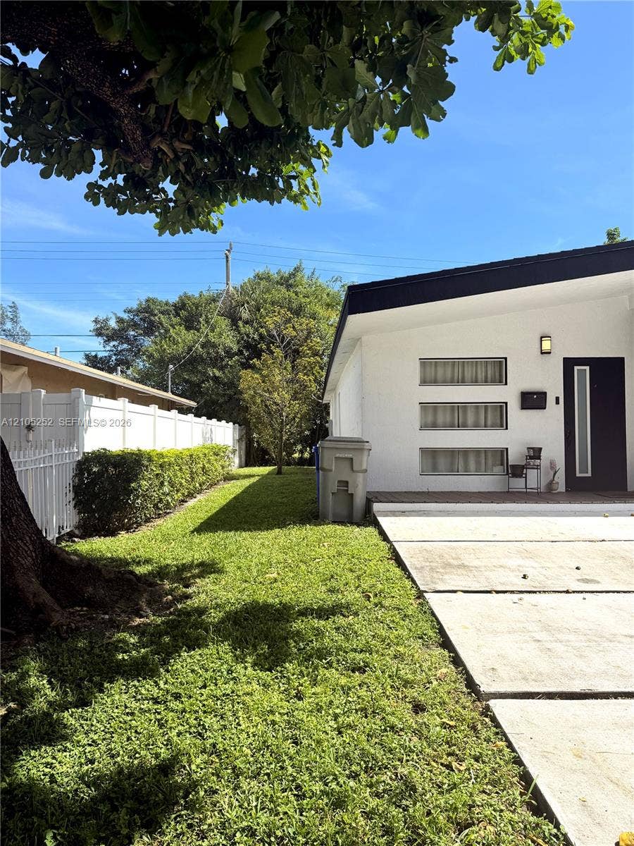 700 NW 17th Terrace, Pompano Beach, FL 33069 White exterior with black trim, concrete walkway, green lawn, white picket fence, and mature landscaping