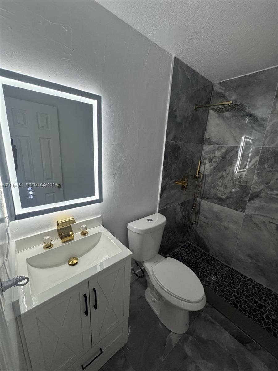 Backlit vanity mirror, white vanity with gold-tone fixtures, dark gray floor tile, dark gray wall tile, and pebble stone shower floor