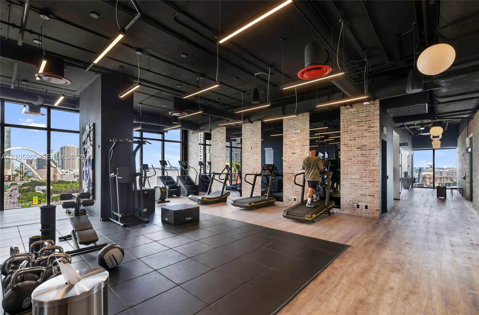 398 NE 5th Street, #2219, Miami, FL 33132 Fitness facility featuring floor-to-ceiling windows, exposed industrial-style black ceilings with integrated lighting, exposed brick accent walls, and dual-tone flooring consisting of square dark tiling and wood-finish planks