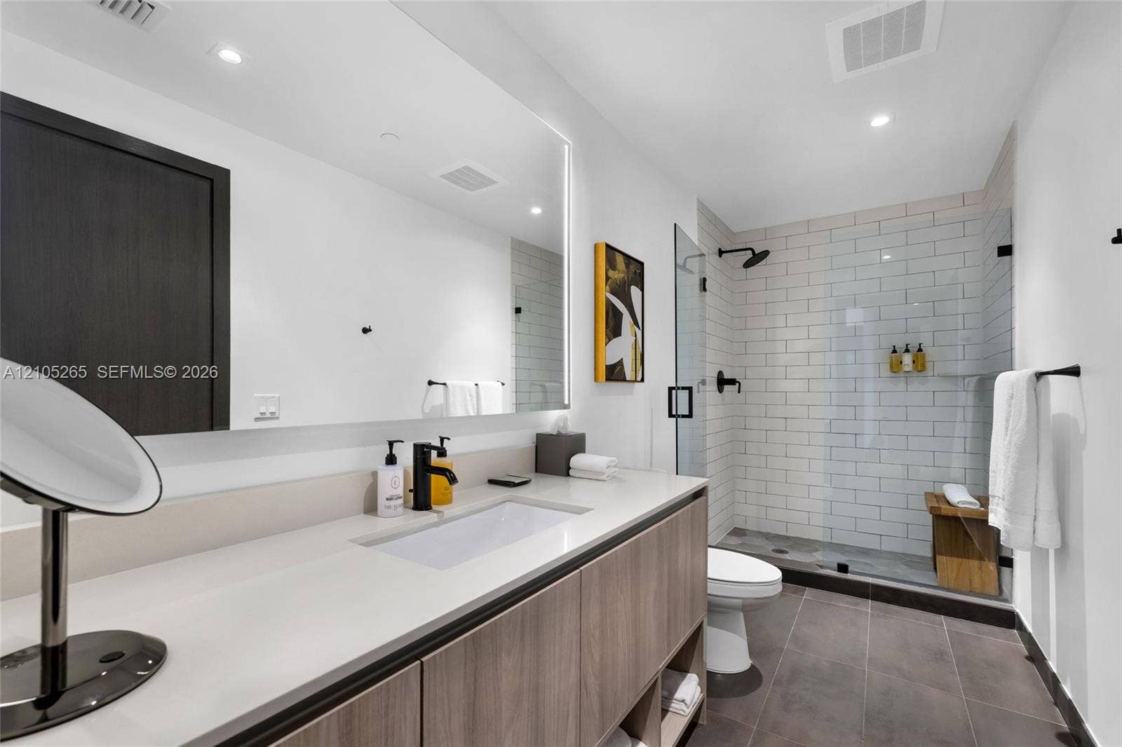 398 NE 5th Street, #2219, Miami, FL 33132 Floating wood-finish vanity with integrated sink and quartz countertop, large backlit mirror, and glass-enclosed shower featuring white subway-style tile and matte black fixtures