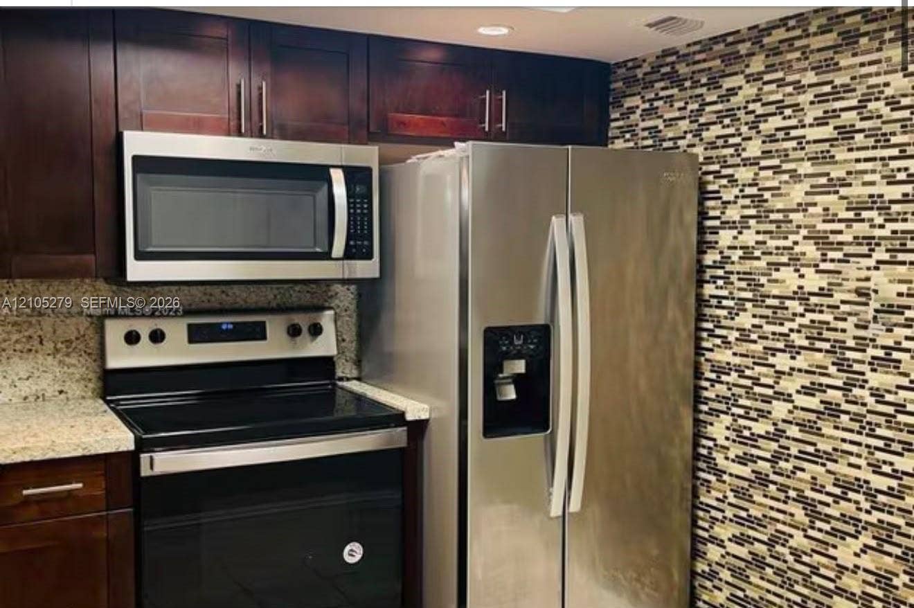 Stainless steel appliances, rich wood-finish cabinetry, patterned mosaic tile backsplash, and speckled countertops